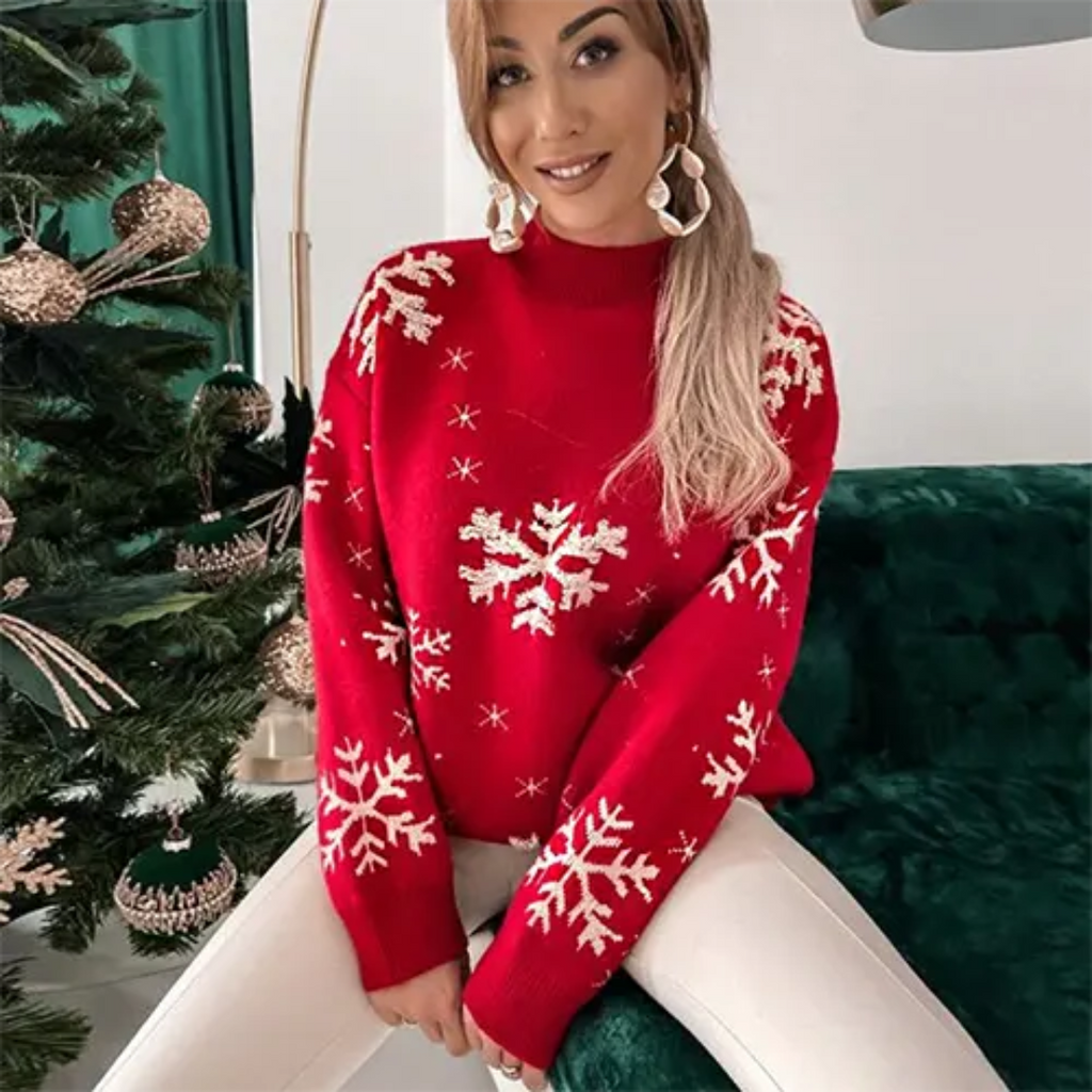 Femme souriante portant un pull rouge à motifs flocons de neige, assise près d'un sapin de Noël décoré. Mode hiver, tenue festive, ambiance Noël.