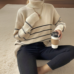 Femme portant un pull à col roulé beige rayé, tenant un café à emporter, assise sur un tapis beige. Mode automne, confort, style décontracté.
