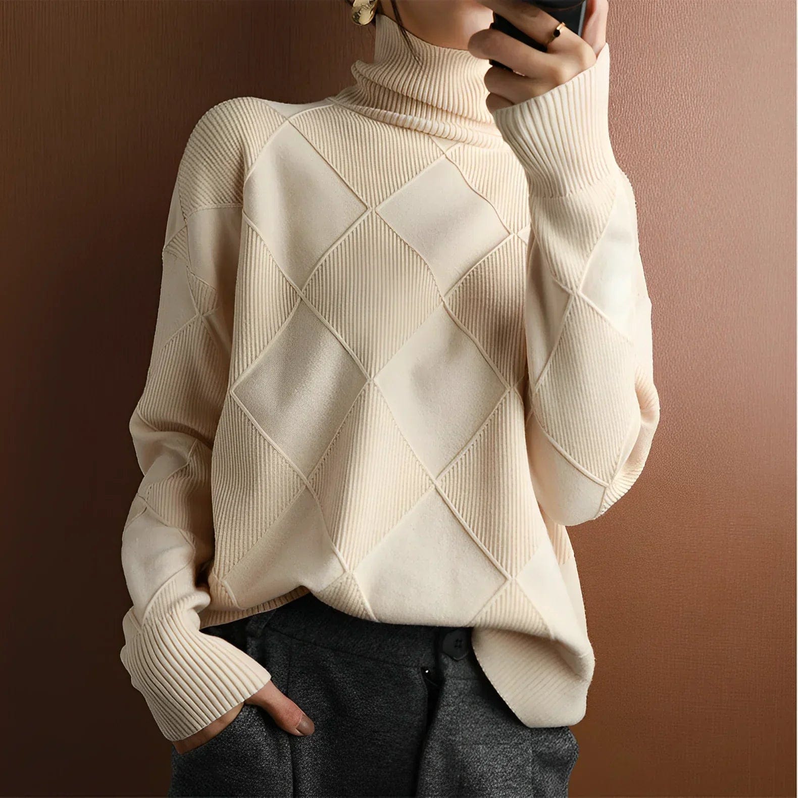 Pull-over beige à col roulé, motif losanges, manches longues, porté par une personne tenant un smartphone. Mode automne-hiver, style élégant et moderne.
