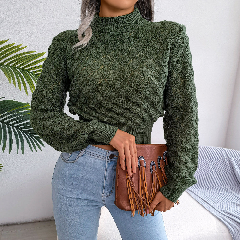 Femme portant un pull vert texturé avec un sac à main marron à franges, posant devant un fond de plantes vertes et un mur blanc.