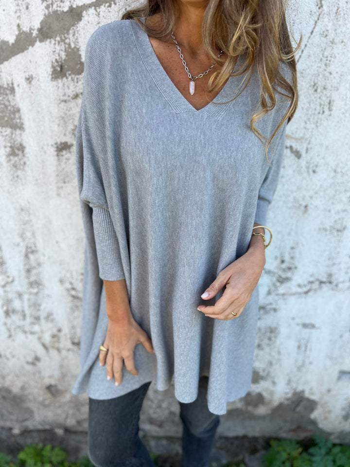 Femme portant un pull gris ample à col en V, posant devant un mur texturé. Mode automne, style décontracté, tendance vêtements confortables.