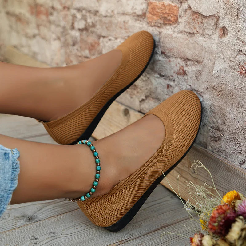 Chaussures plates marron en tissu côtelé, portées avec un jean déchiré et un bracelet turquoise, posées sur un sol en bois devant un mur de briques.