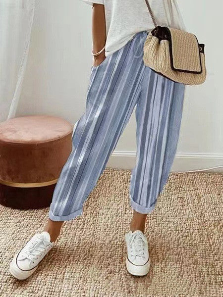 Femme portant un pantalon rayé bleu et blanc, t-shirt blanc, baskets blanches et sac en osier. Mode décontractée, style estival, intérieur moderne.