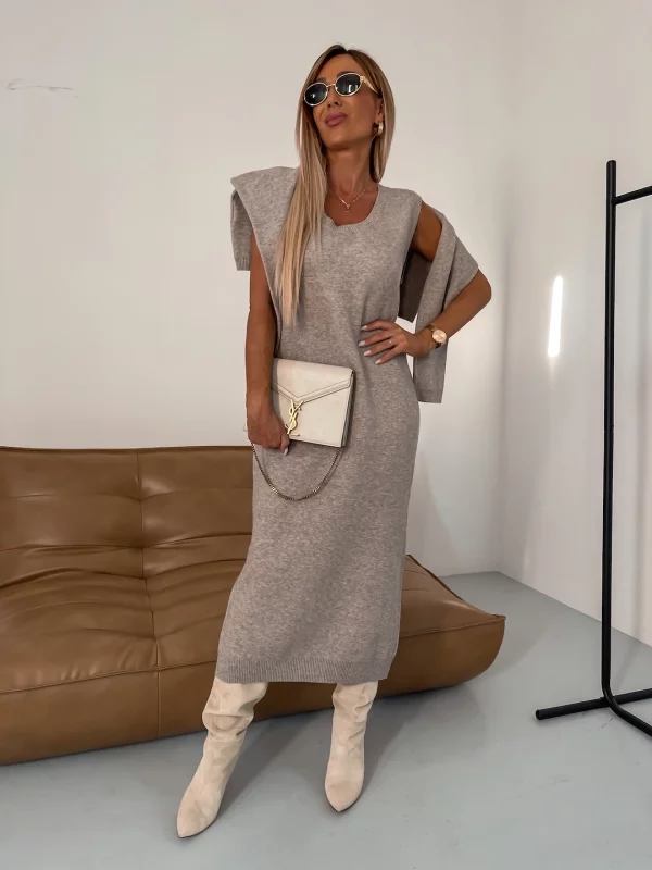 Femme élégante en robe pull grise, portant des bottes blanches et lunettes de soleil, posant dans un salon moderne avec canapé en cuir marron.
