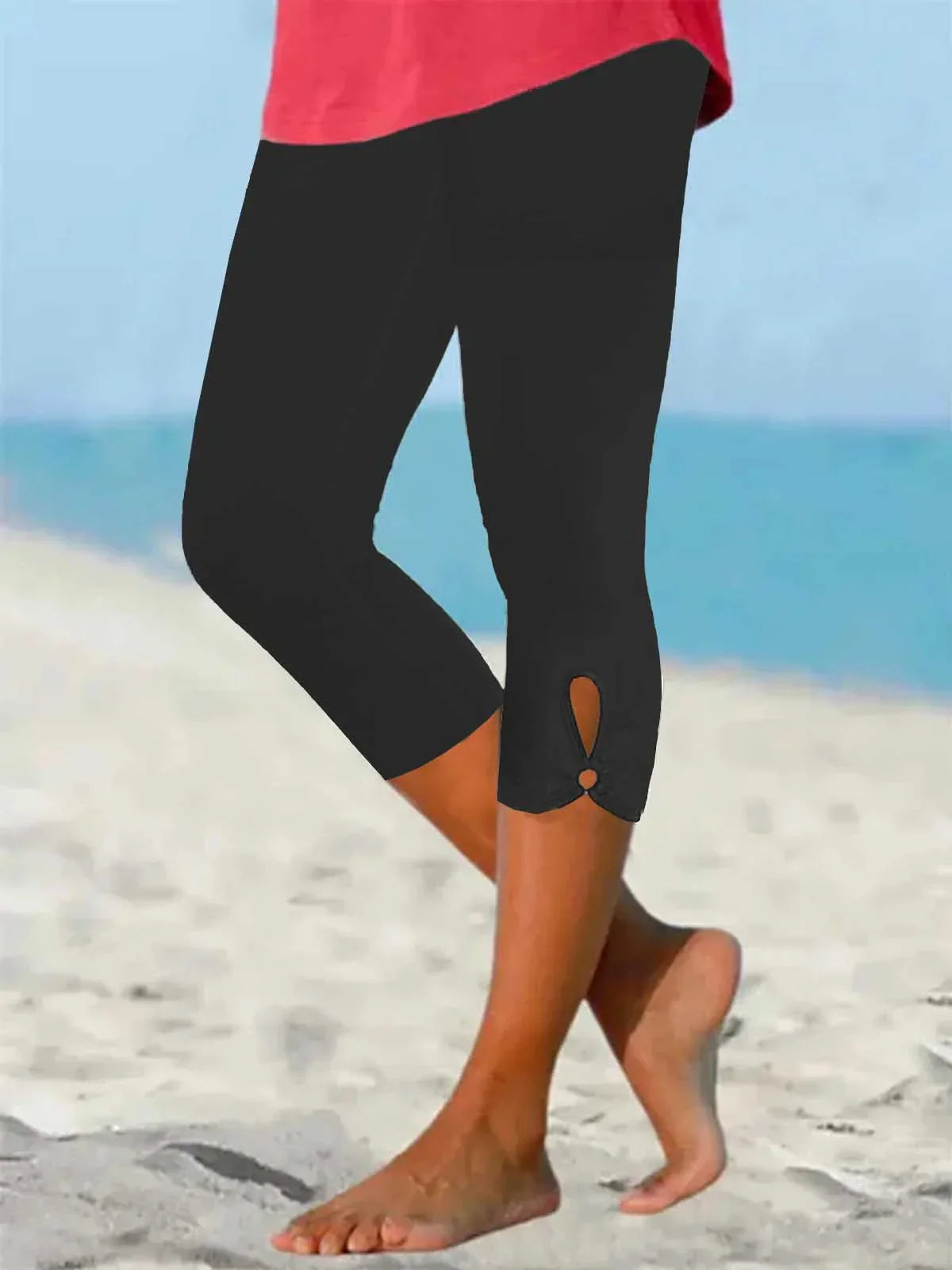 Femme portant un legging noir 3/4 avec détail découpé, marchant pieds nus sur une plage. Mode estivale, confort, style décontracté.
