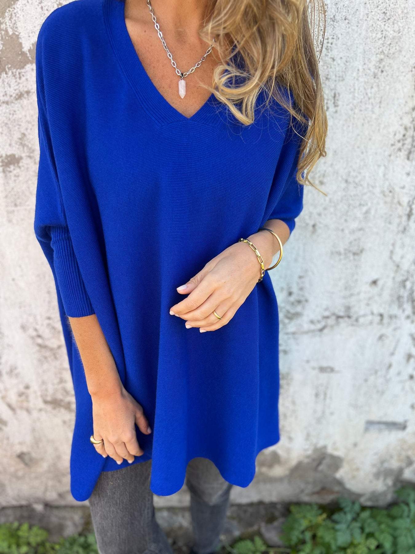 Femme portant un pull bleu vif, manches longues, col en V, avec un collier argenté et un bracelet doré, posant devant un mur texturé.