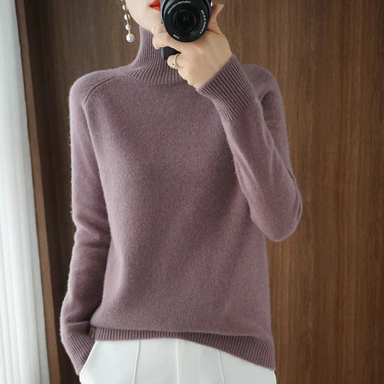 Femme en pull col roulé mauve, tenant un appareil photo, devant un fond en bois. Mode automne-hiver, style élégant et décontracté.