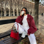 Femme assise sur un banc, portant une veste rouge et buvant du café. Sac à main rouge et baskets blanches. Arbres dénudés en arrière-plan.