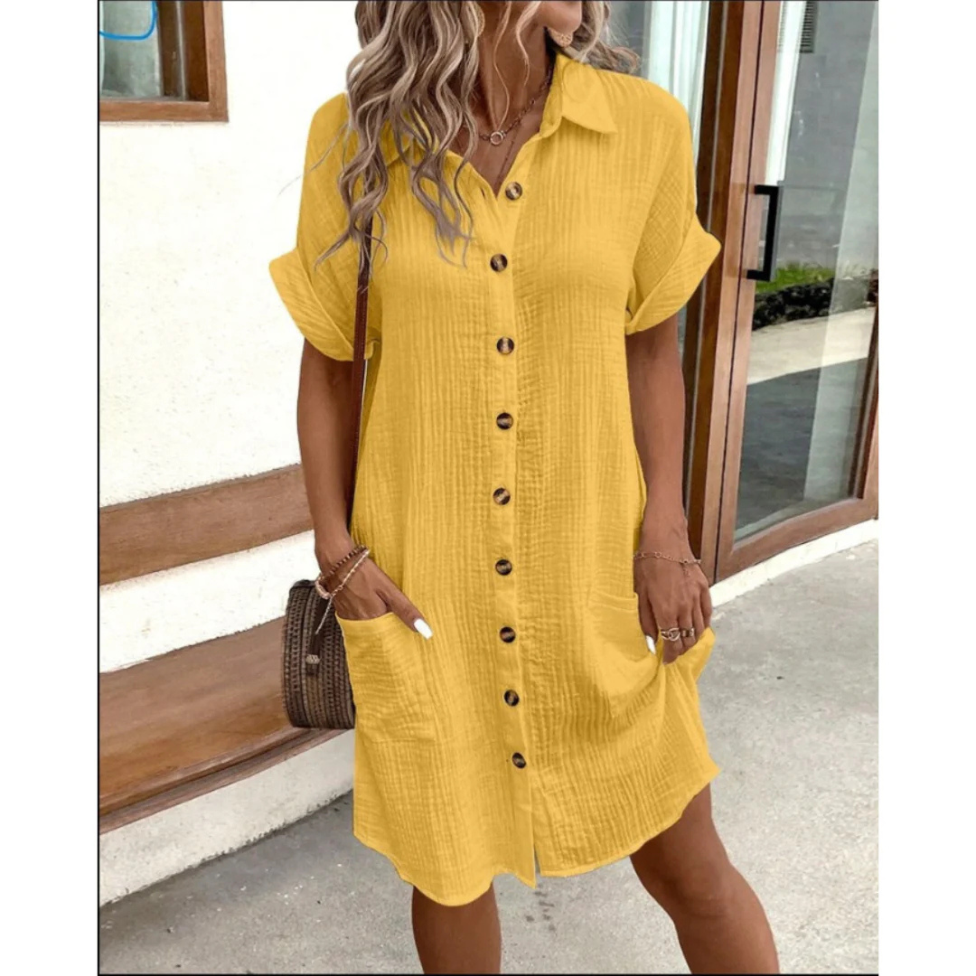 Robe chemise jaune à manches courtes, boutons en bois, poches latérales, portée par une femme aux cheveux ondulés, devant une porte vitrée.