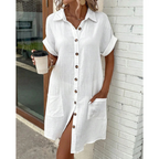 Robe chemise blanche en lin avec boutons en bois, manches courtes retroussées, poches avant, femme tenant un café devant un mur de briques blanches.