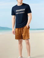 Homme en t-shirt bleu marine avec texte blanc et short marron sur une plage ensoleillée. Mode estivale, tenue décontractée, bord de mer.