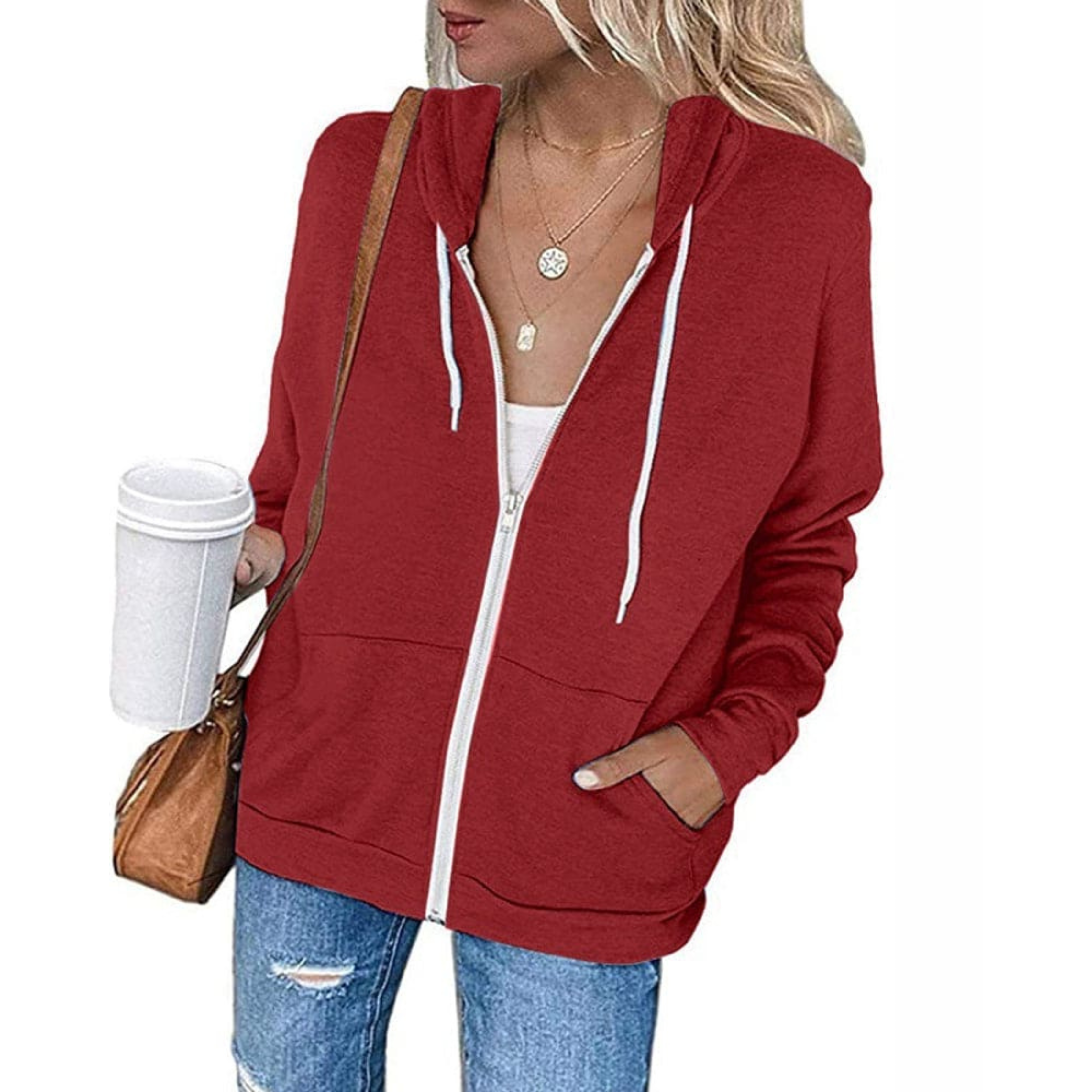 Femme portant un sweat à capuche rouge avec fermeture éclair, tenant un gobelet à café, sac en cuir marron, collier doré, jeans décontracté.