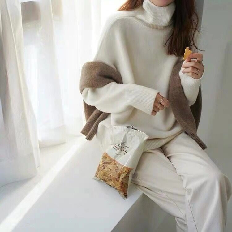 Femme en pull blanc et pantalon beige mangeant des chips près d'une fenêtre. Mode automne, col roulé, détente, collation.