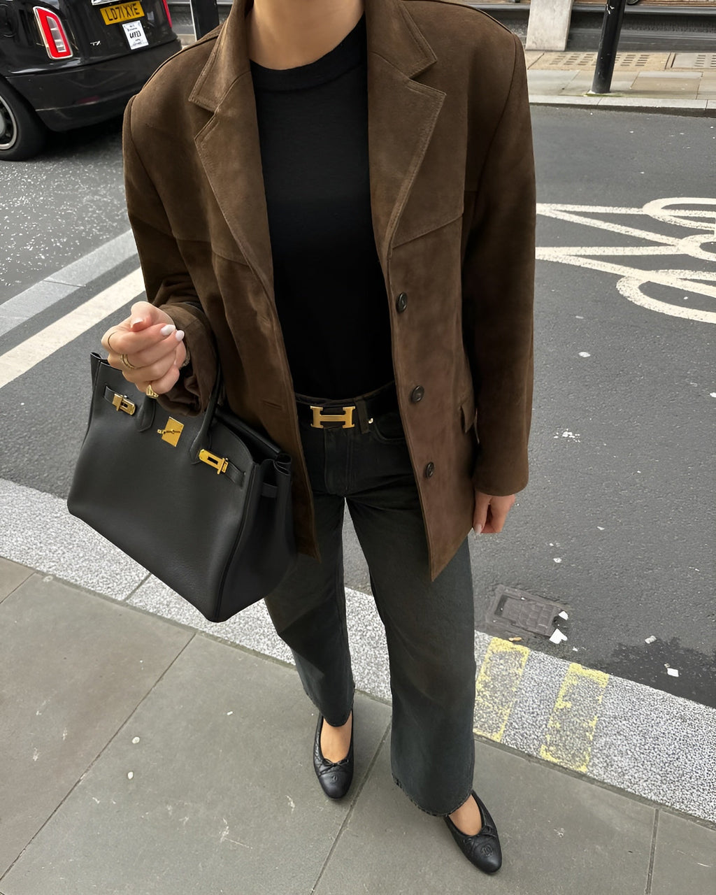 Femme en veste marron, jean noir, ceinture Hermès, sac Birkin noir, chaussures noires, sur trottoir urbain. Mode élégante et chic.