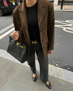 Femme en veste marron, jean noir, ceinture Hermès, sac Birkin noir, chaussures noires, sur trottoir urbain. Mode élégante et chic.