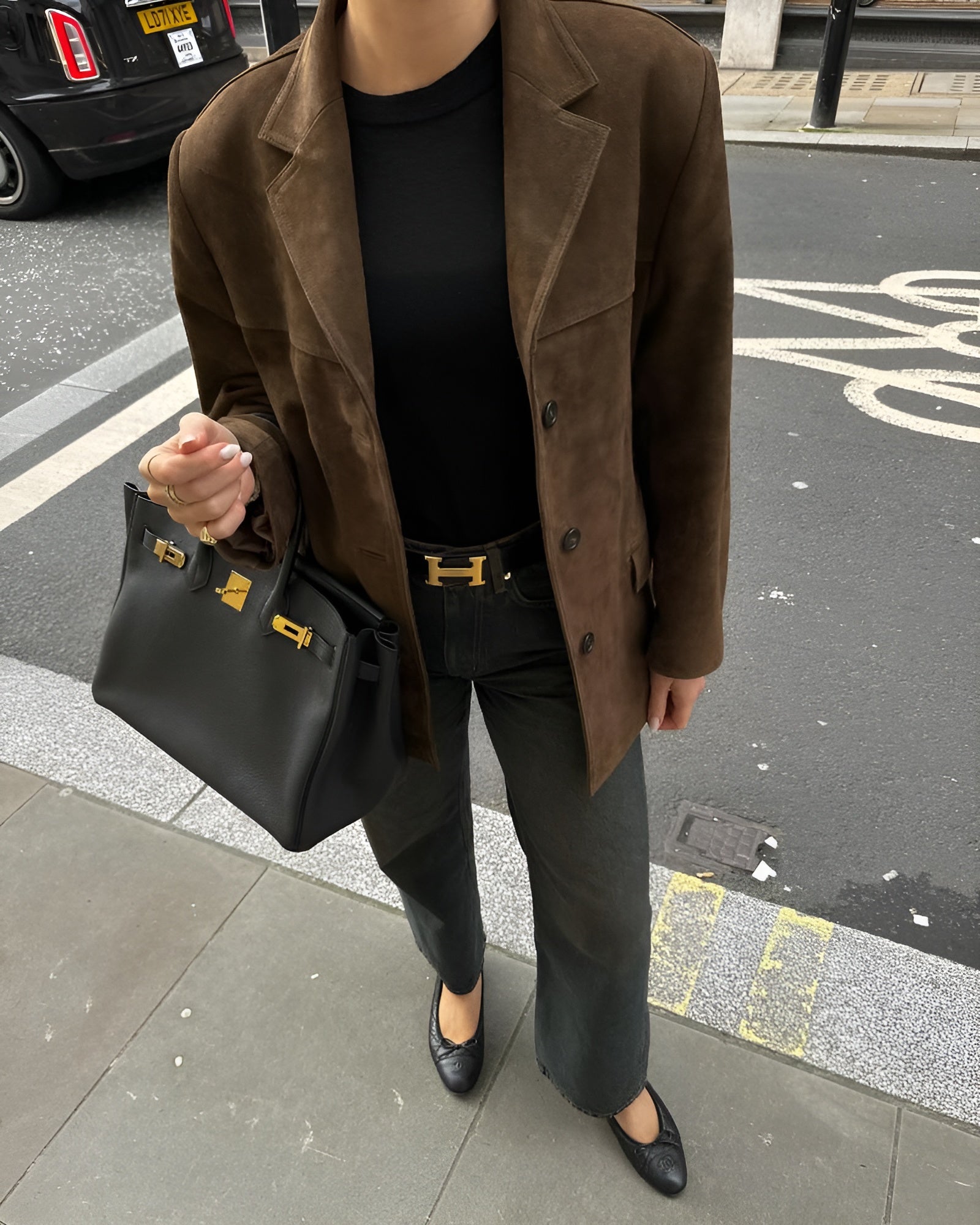 Femme en veste marron, jean noir, ceinture Hermès, sac Birkin noir, chaussures noires, sur trottoir urbain. Mode élégante et chic.