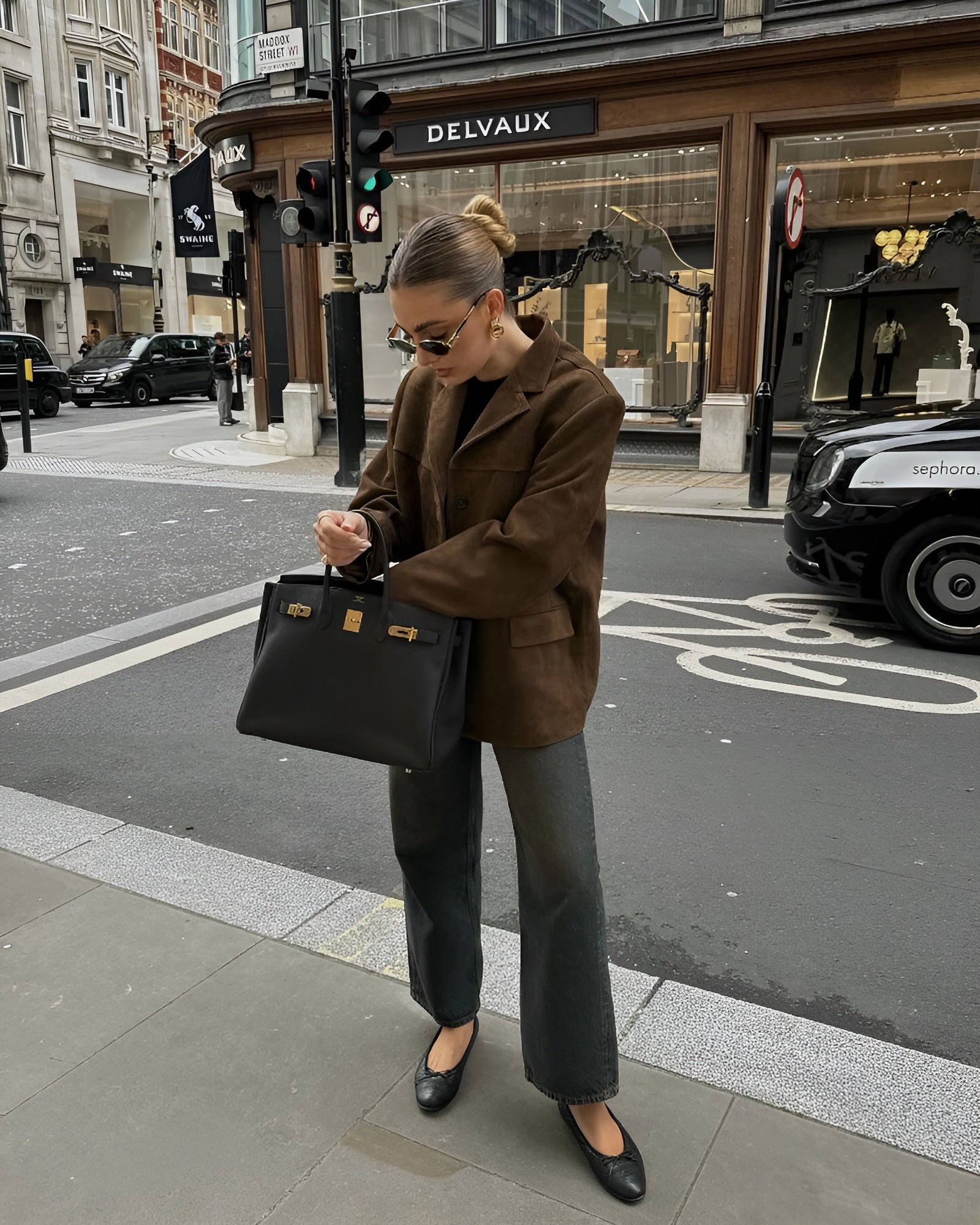 Femme élégante portant un manteau marron et un sac à main noir devant une boutique Delvaux à Londres, rue animée avec taxis et piétons.