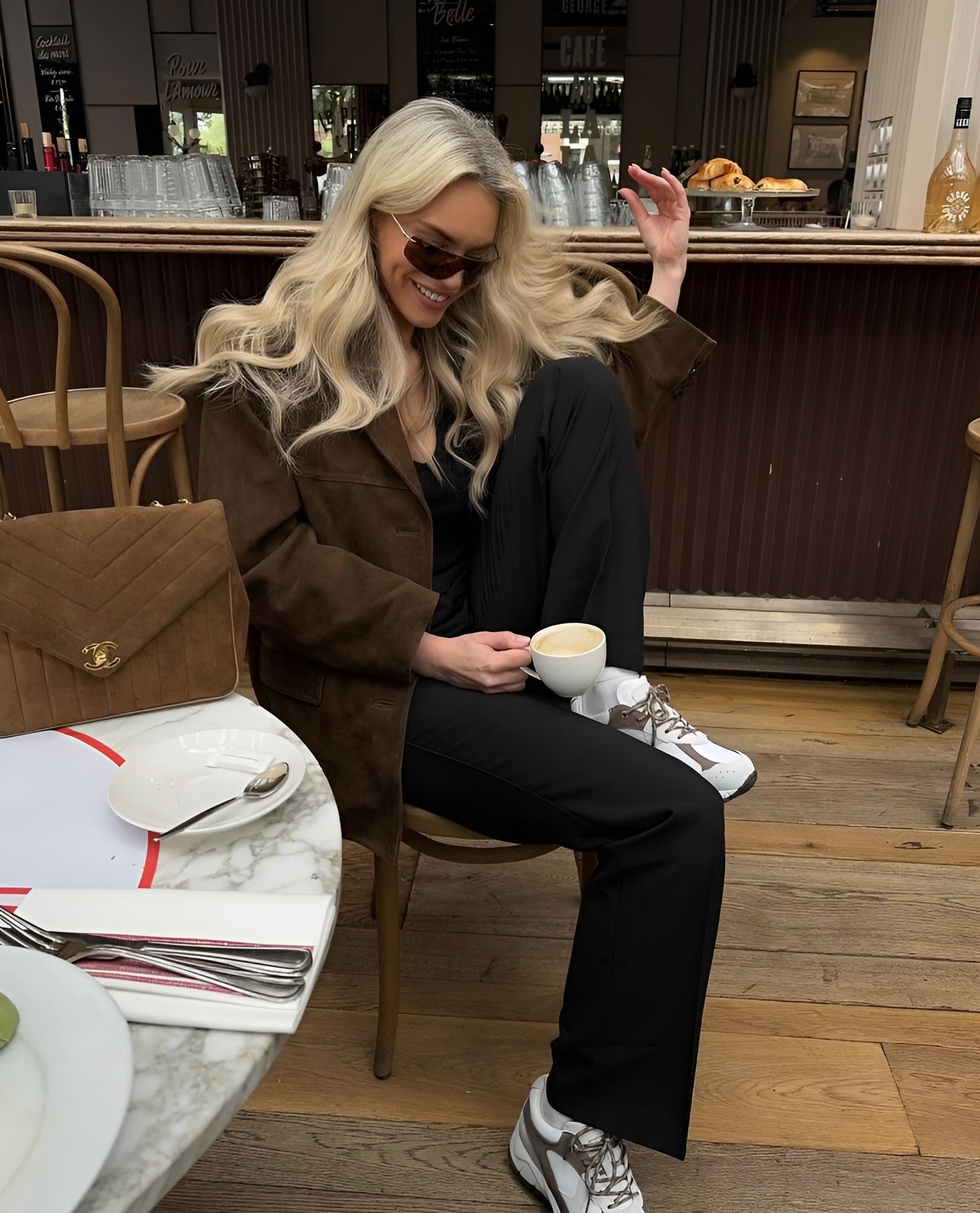 Femme blonde en lunettes de soleil, veste marron, pantalon noir, assise dans un café avec une tasse de café. Sac à main et assiette sur la table.