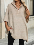 Femme portant un pull beige à col zippé, manches courtes, avec un collier doré, posant devant un bâtiment moderne. Mode automne chic et décontractée.