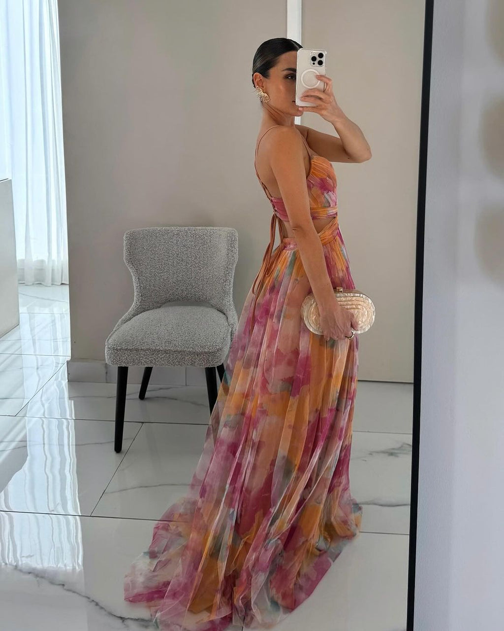 Robe longue élégante à motifs floraux colorés, femme prenant un selfie dans un miroir, tenue de soirée chic, sac à main doré, intérieur moderne.