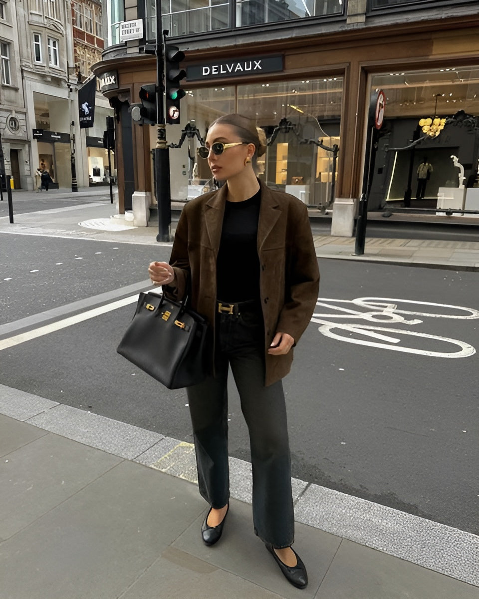 Femme élégante en manteau marron et lunettes de soleil, tenant un sac à main noir Hermès, traverse une rue urbaine devant une boutique Delvaux.