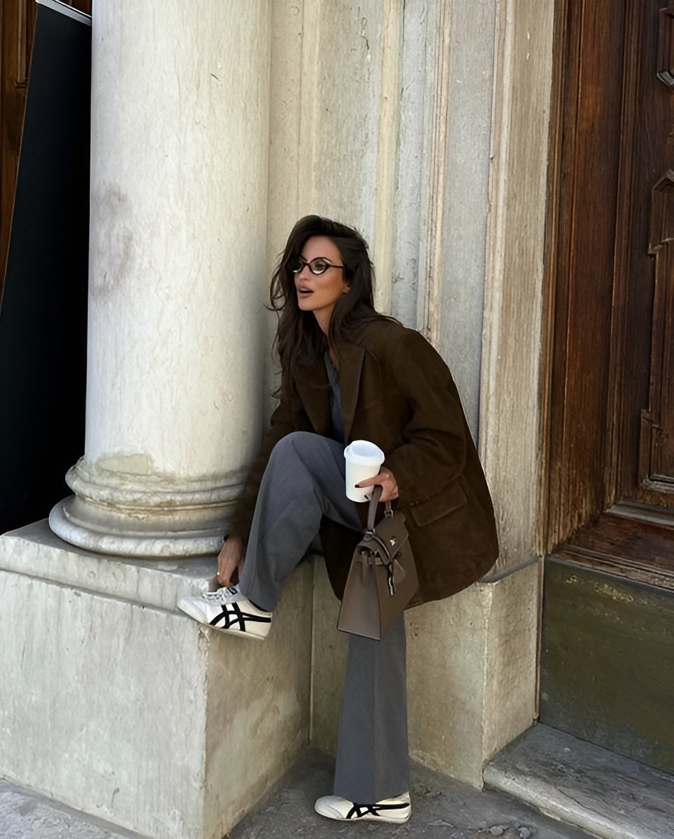 Femme élégante en manteau marron, pantalon gris, baskets blanches, tenant un café et un sac à main, posant contre une colonne en pierre.