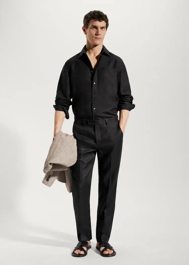 Homme en chemise noire et pantalon noir, tenant une veste beige. Mode masculine élégante, tenue décontractée chic, style moderne.