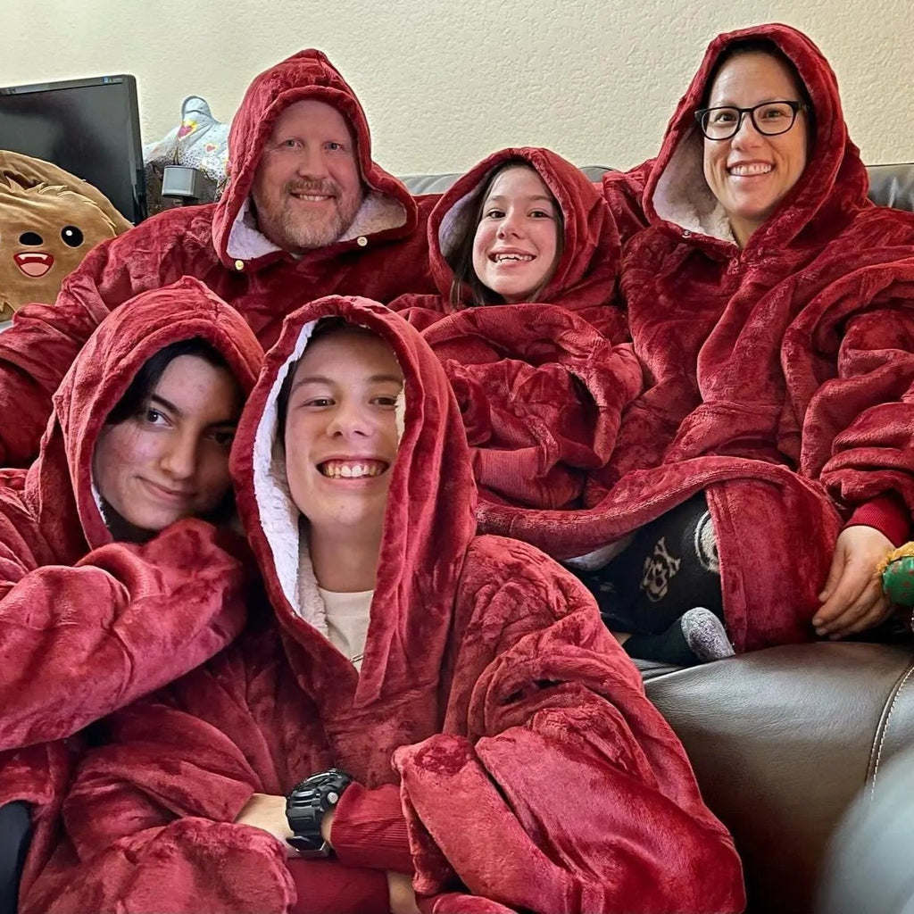 Cinq personnes souriantes portant des couvertures à capuche rouges, assises sur un canapé. Ambiance chaleureuse et conviviale.
