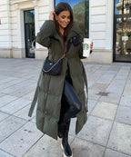 Femme en manteau long vert, leggings en cuir noir, tenant un café Starbucks, devant un bâtiment blanc. Mode hiver, style urbain chic.