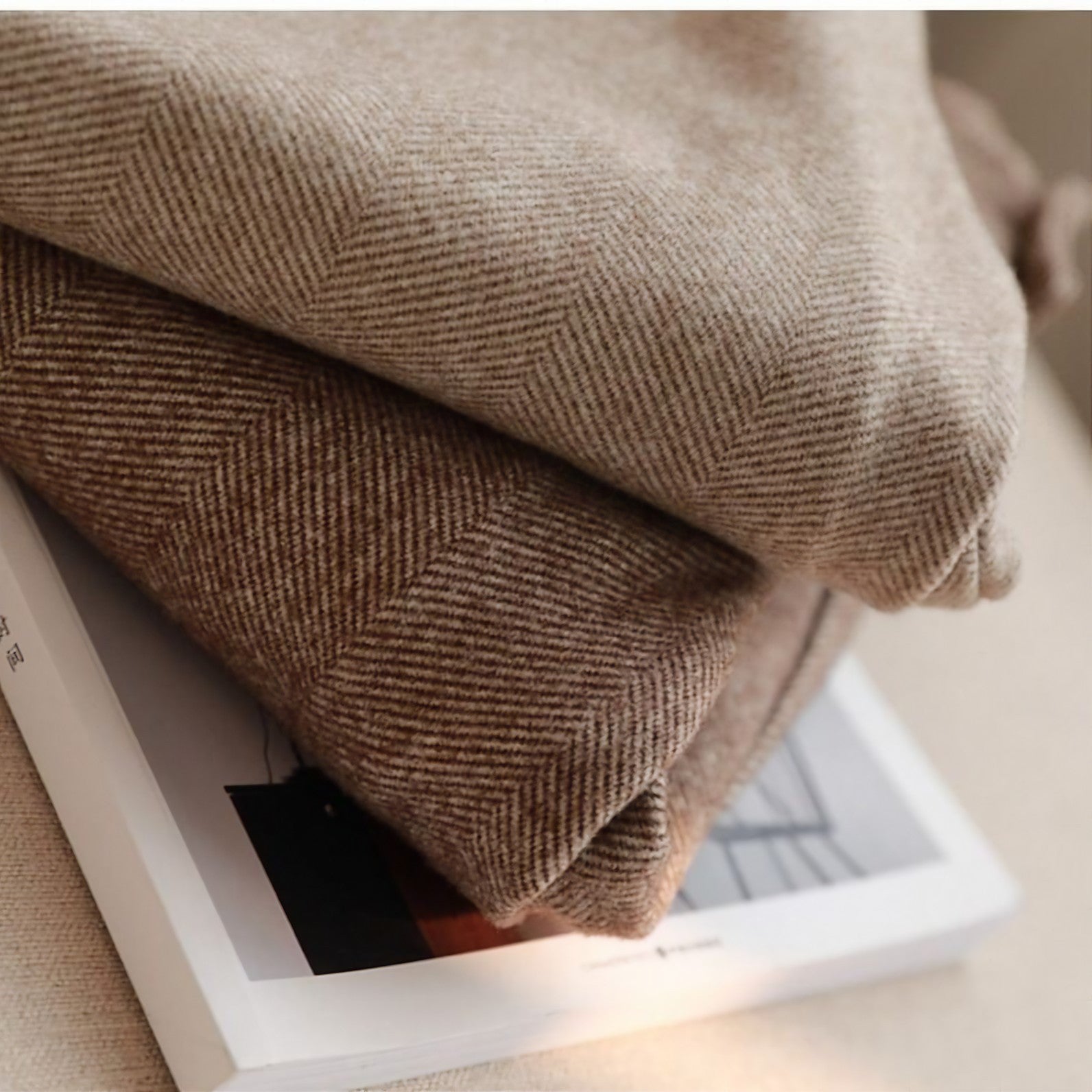 Deux couvertures en laine beige et marron à motif chevron, posées sur un livre, créant une ambiance cosy et chaleureuse.