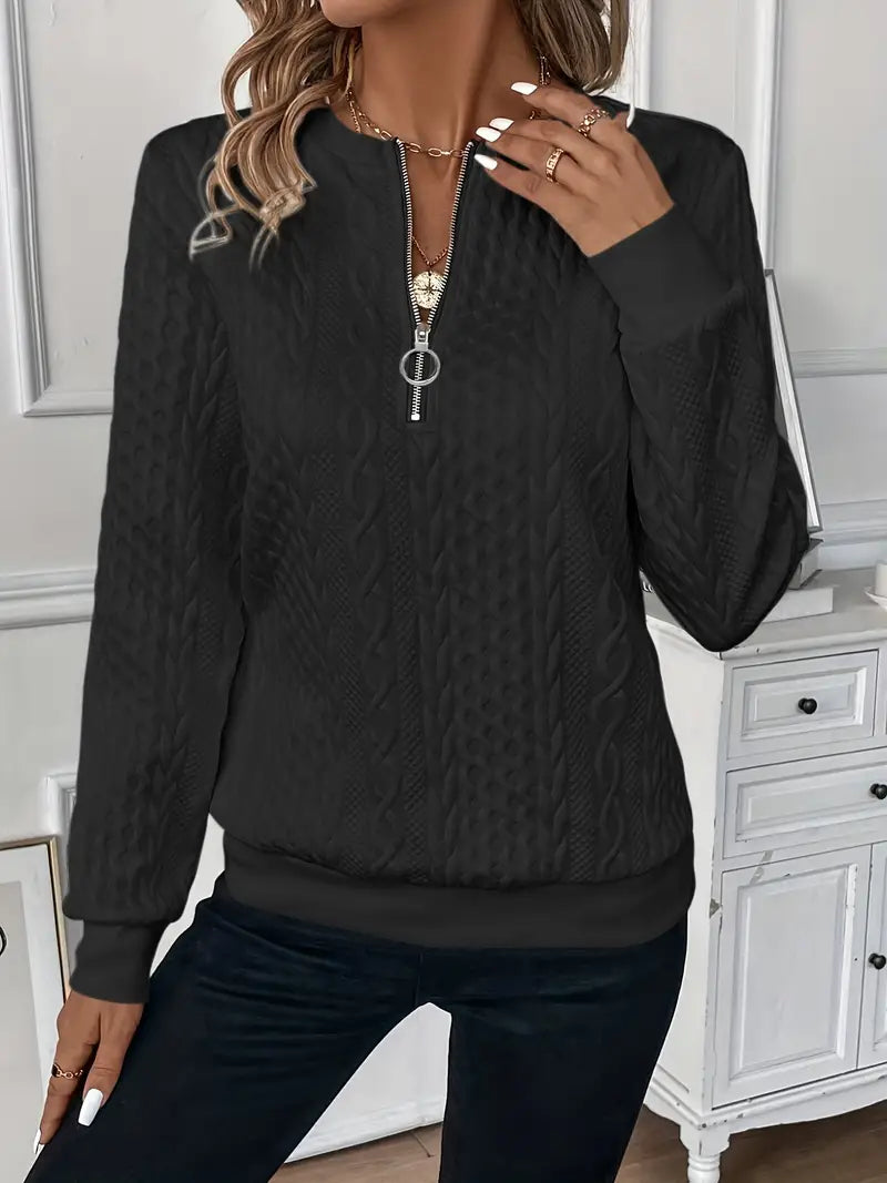 Femme portant un pull noir à fermeture éclair, motif torsadé, manches longues, devant un meuble blanc. Mode automne-hiver, style décontracté.