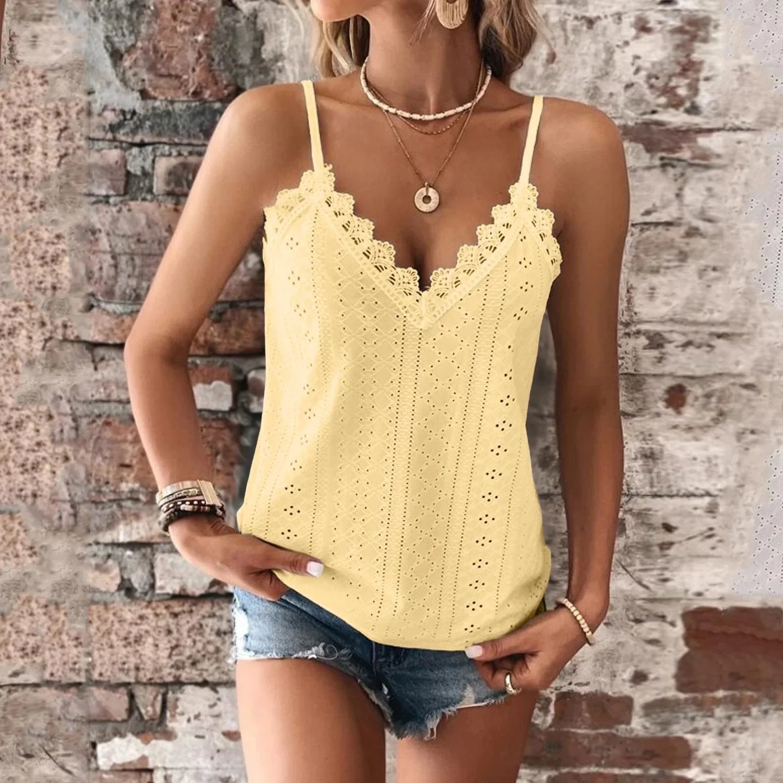 Femme portant un débardeur jaune en dentelle avec short en jean, posant devant un mur en briques. Mode estivale, tenue décontractée, accessoires bohèmes.