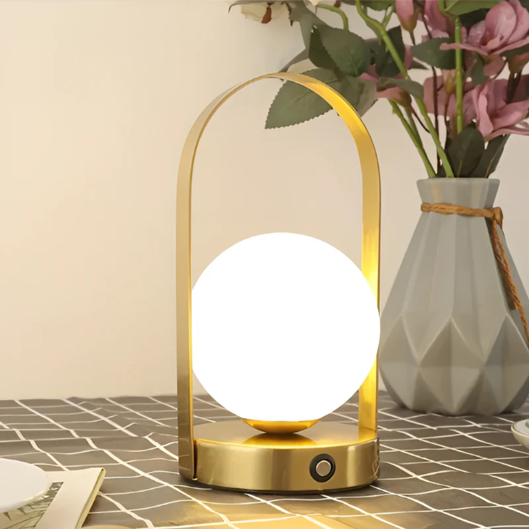 Lampe de table moderne en métal doré avec globe lumineux, posée sur une table à carreaux, vase gris avec fleurs en arrière-plan. Éclairage design élégant.