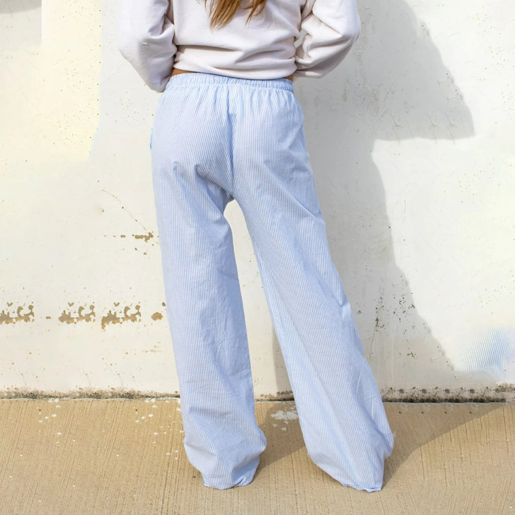 Femme portant un pantalon ample bleu clair à rayures, debout contre un mur beige. Mode décontractée, style confortable, tendance féminine.
