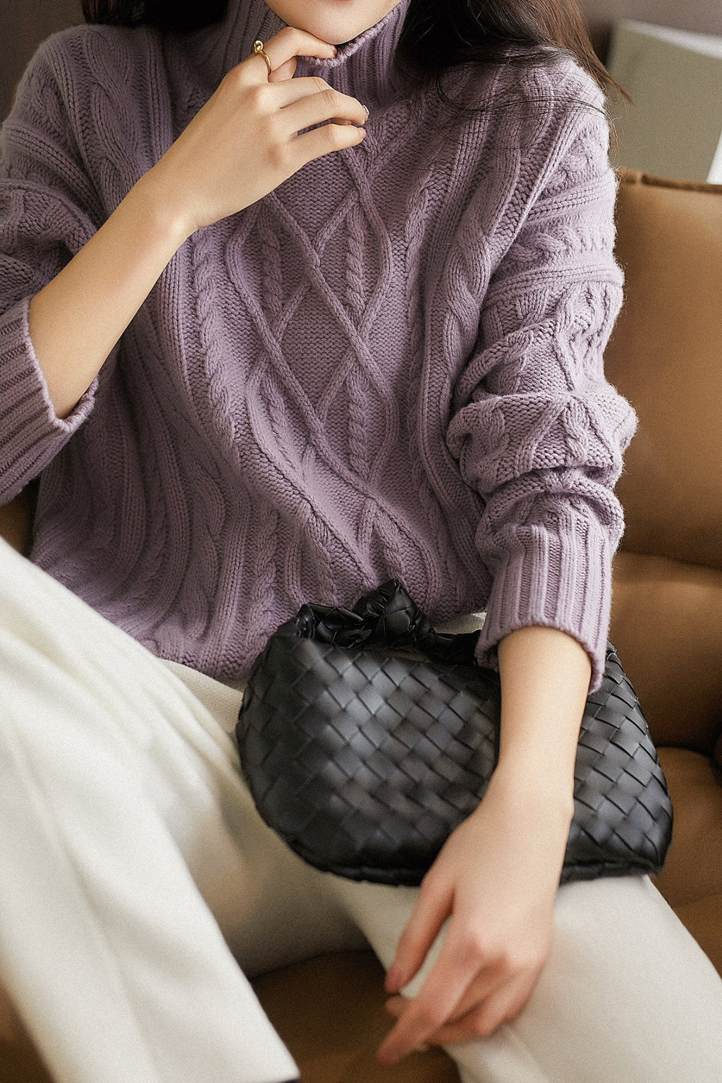 Femme en pull violet torsadé, tenant un sac à main noir tressé, assise sur un canapé marron. Mode automne, style élégant et décontracté.
