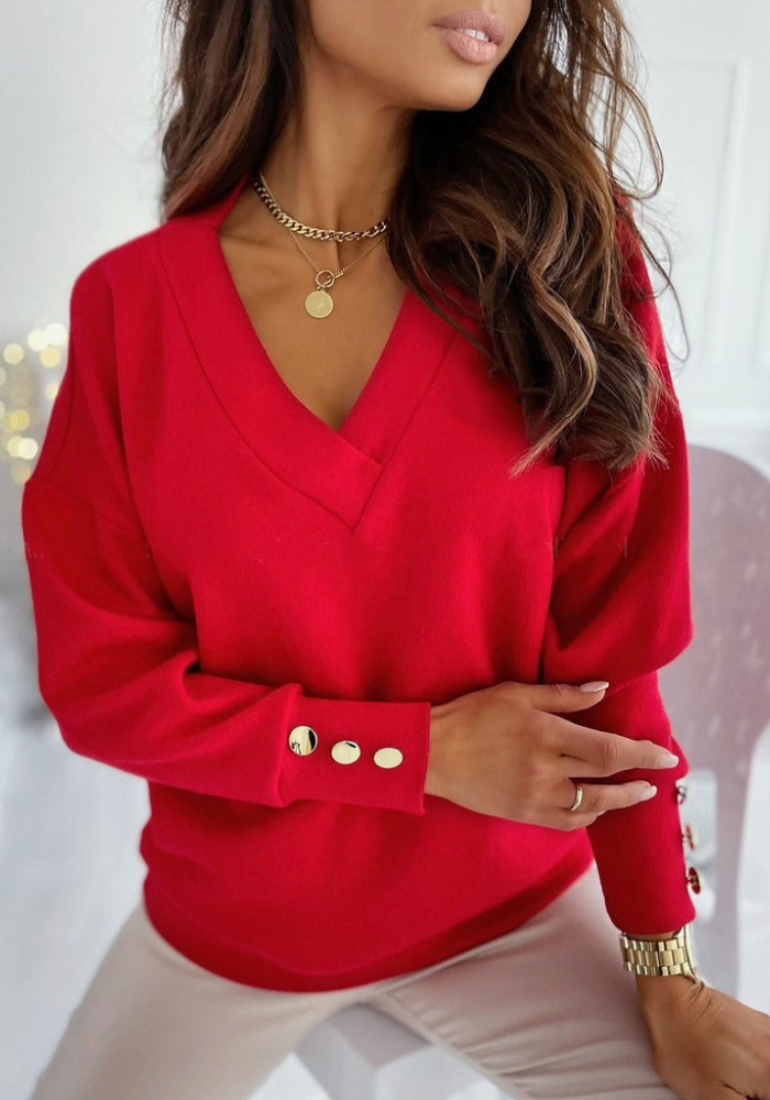 Femme portant un pull rouge à col en V avec boutons dorés, collier doré, posant élégamment. Mode automne-hiver, style chic et tendance.