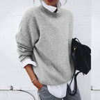 Femme portant un pull gris ample sur une chemise blanche, tenant un sac noir. Mode décontractée élégante, tenue automne-hiver tendance.