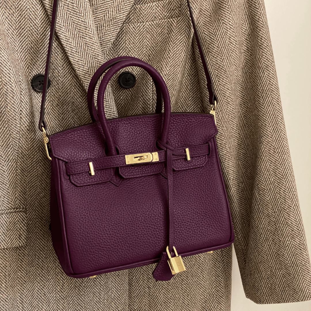 Sac à main en cuir violet avec fermoir doré, suspendu à un manteau en tweed marron. Accessoire de mode élégant et tendance.