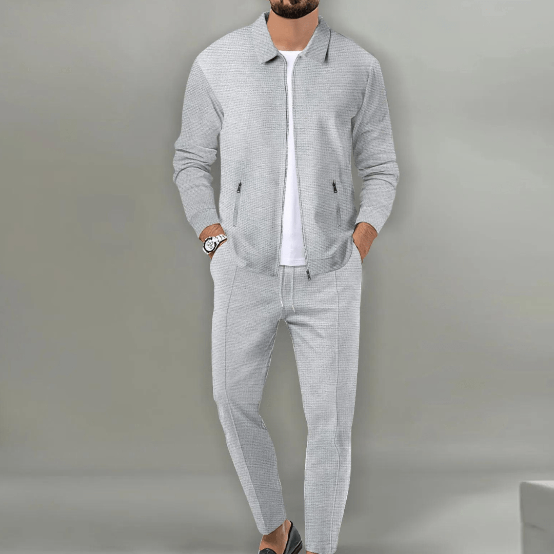 Homme portant un ensemble décontracté gris clair, veste et pantalon assortis, sur fond neutre. Mode masculine élégante et moderne.