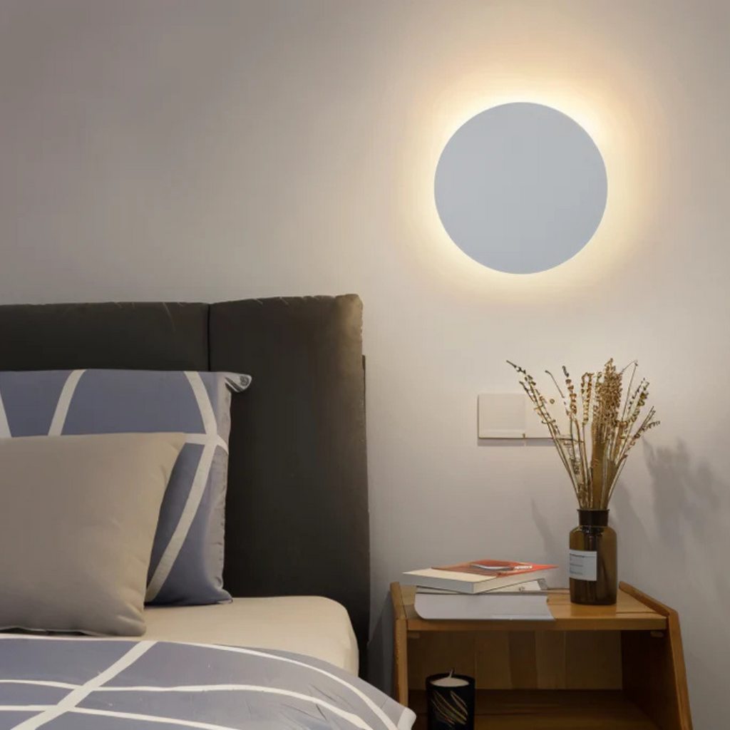 Lampe murale LED ronde moderne éclairant une chambre minimaliste avec lit gris, coussins géométriques, table de chevet en bois et vase décoratif.