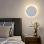 Lampe murale LED ronde moderne éclairant une chambre minimaliste avec lit gris, coussins géométriques, table de chevet en bois et vase décoratif.