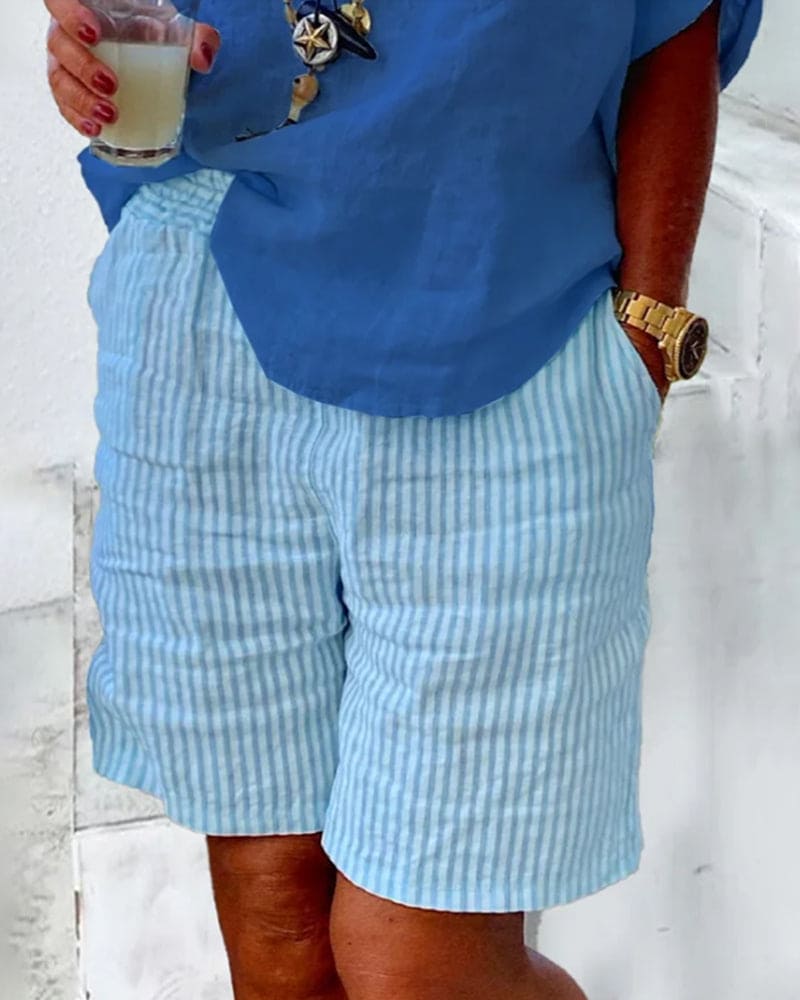 Femme portant un short rayé bleu et blanc, chemise bleue, montre dorée, tenant un verre. Mode estivale décontractée, tenue élégante et confortable.