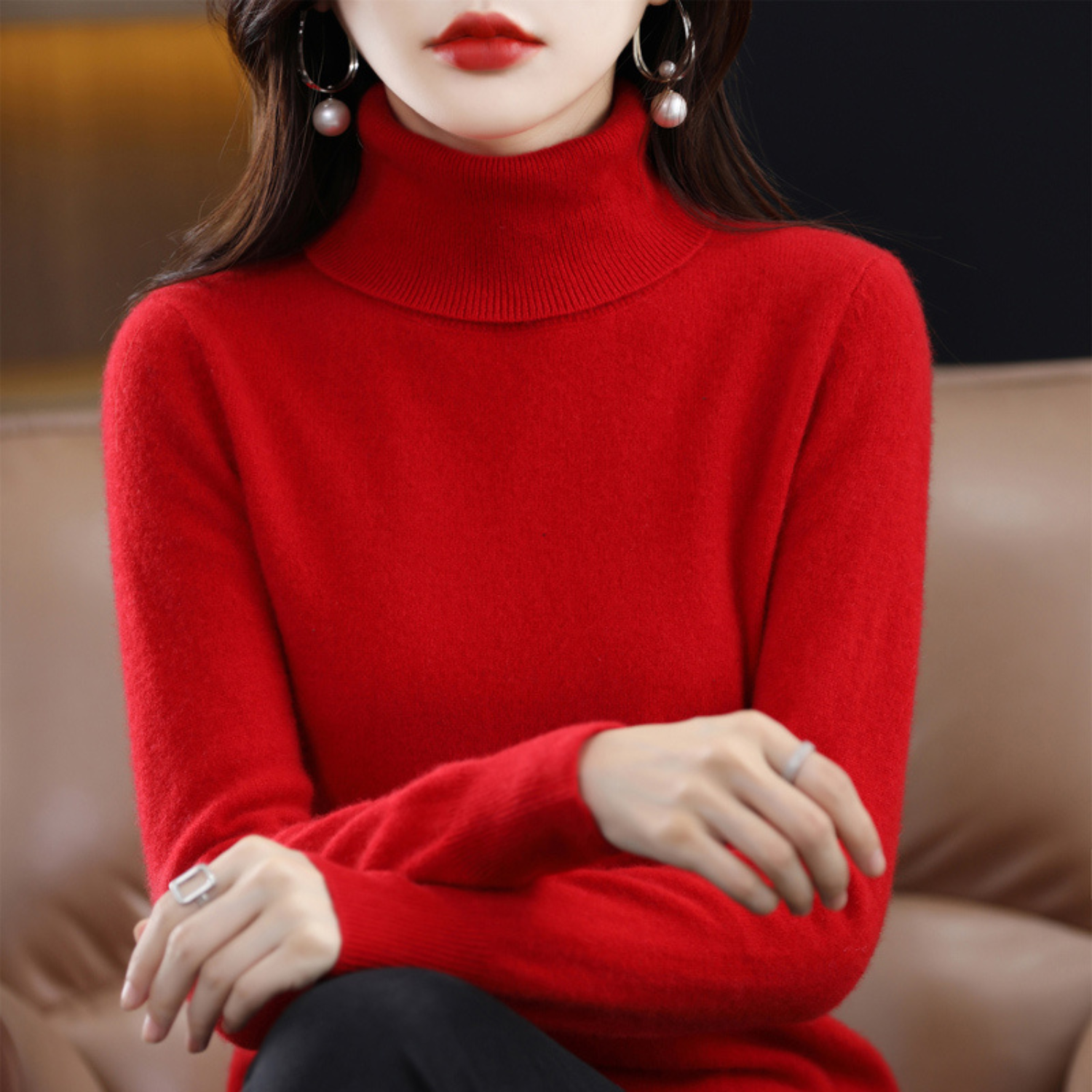 Femme en pull rouge à col roulé, assise sur un canapé en cuir, portant des boucles d'oreilles perle et rouge à lèvres assorti. Mode élégante automne-hiver.