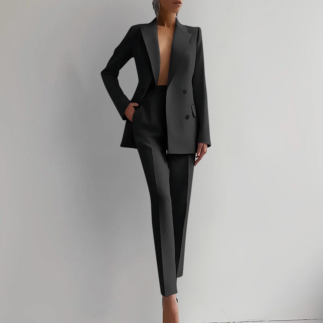 Femme en tailleur noir élégant, veste ouverte, posant contre un mur blanc. Mode féminine, style chic, tenue professionnelle moderne.