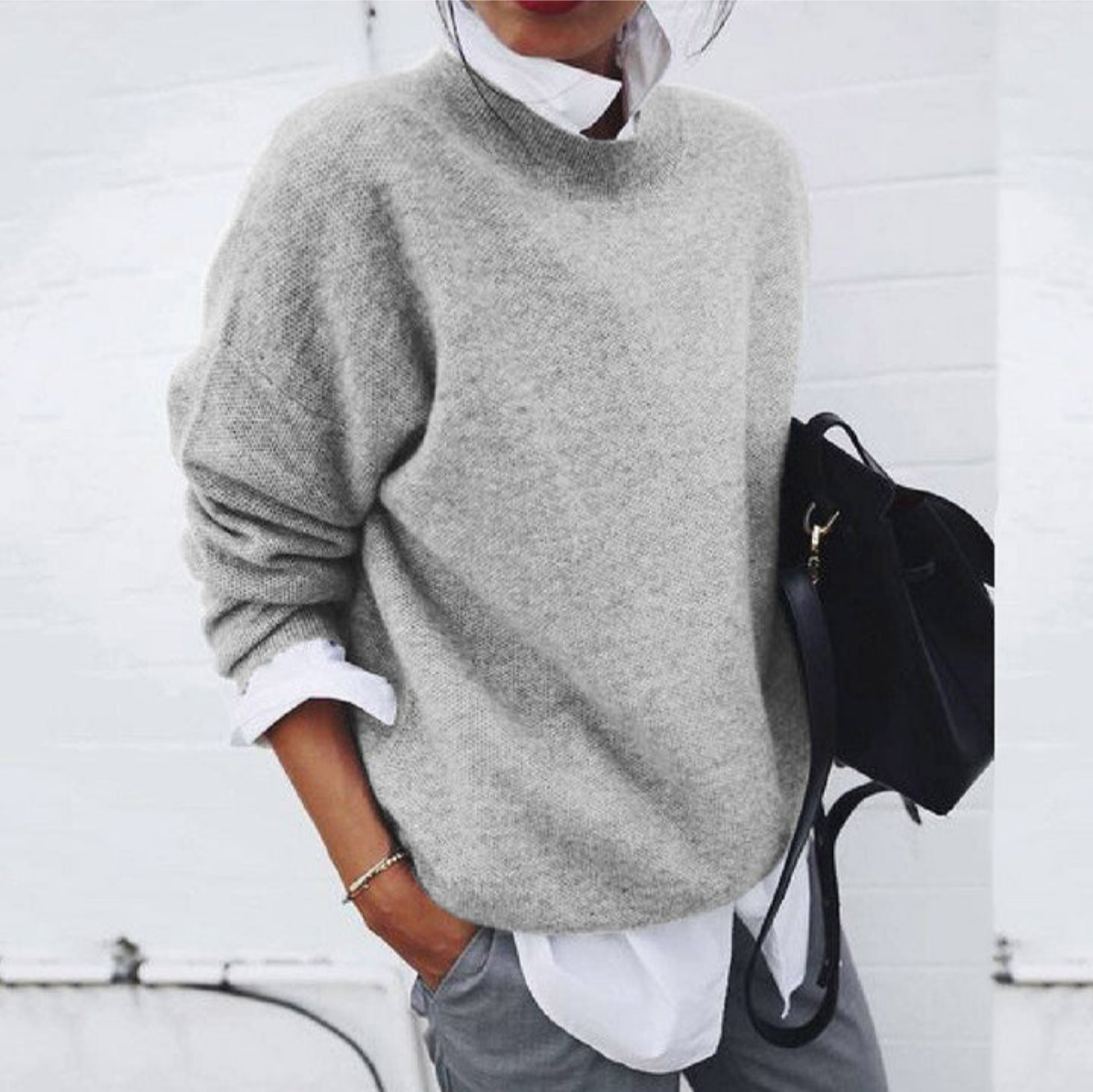 Femme portant un pull gris ample sur une chemise blanche, tenant un sac noir. Mode décontractée chic, style automne-hiver tendance.