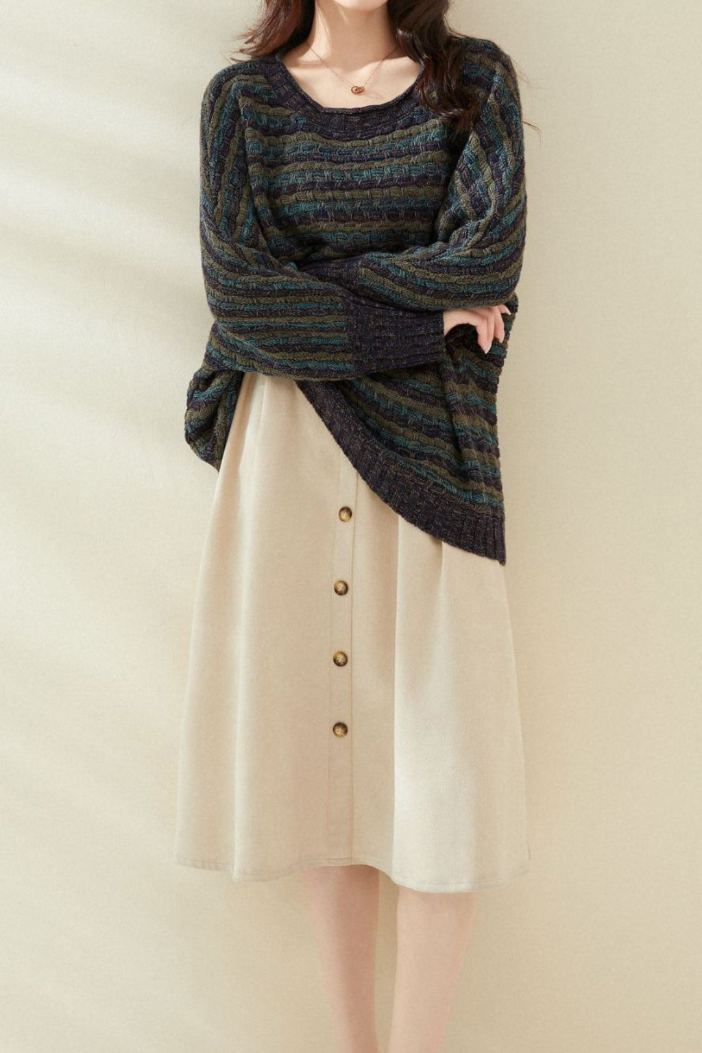 Femme portant une robe beige boutonnée et un pull rayé multicolore, posant contre un mur beige. Mode automne-hiver, tenue décontractée élégante.
