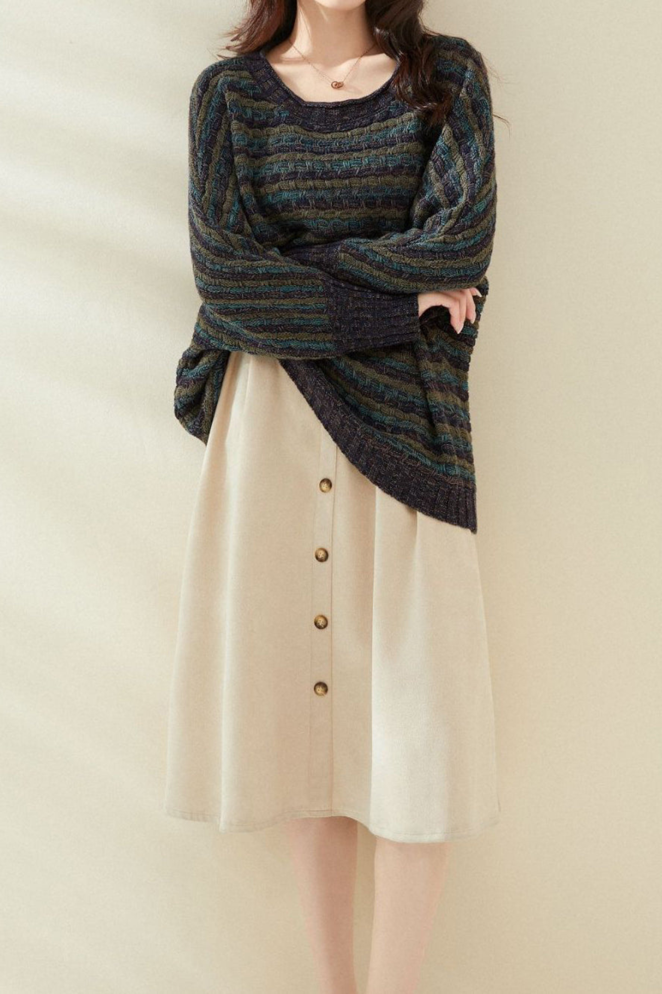 Femme portant une robe beige boutonnée et un pull rayé multicolore, posant contre un mur beige. Mode automne-hiver, tenue décontractée élégante.