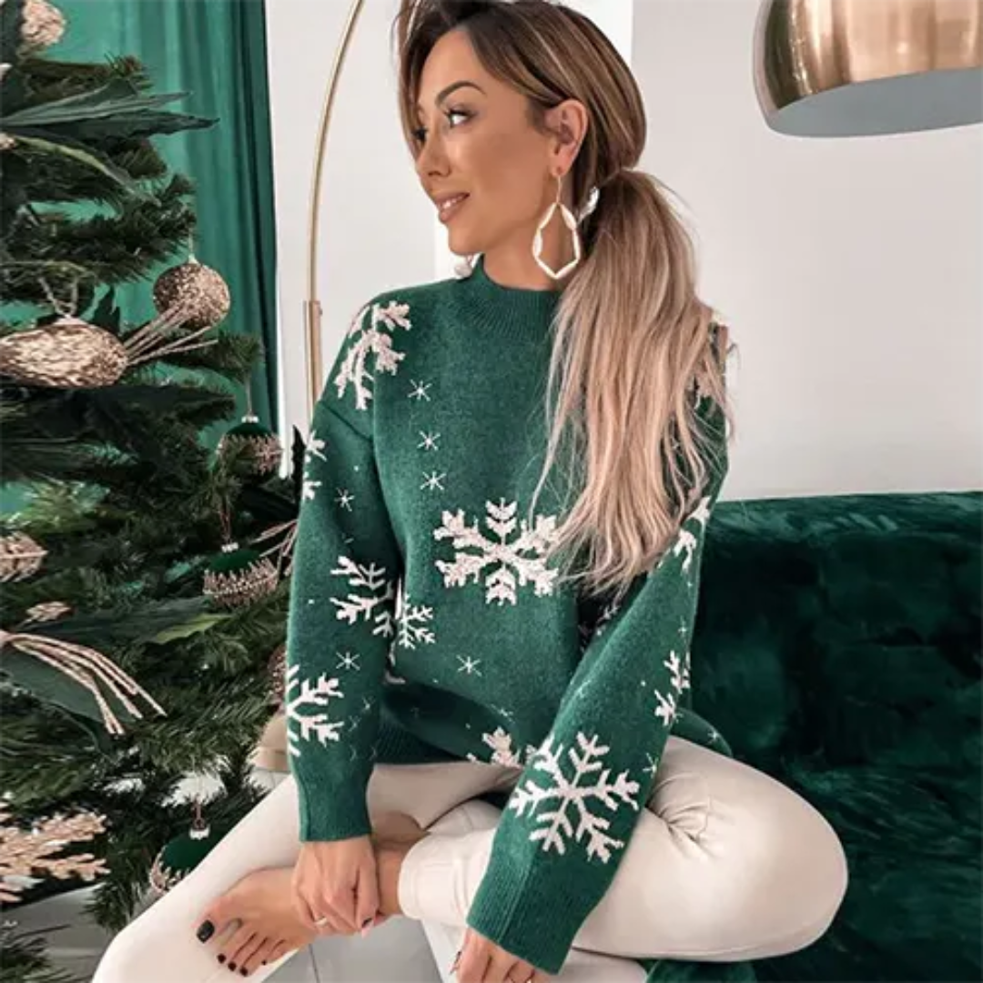 Femme portant un pull vert à motifs flocons de neige, assise près d'un sapin de Noël décoré. Mode hiver, tenue festive, ambiance Noël.