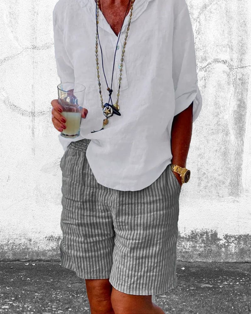 Homme en chemise blanche et short rayé, tenant un verre de boisson. Style décontracté, accessoires tendance, montre dorée. Mode estivale.