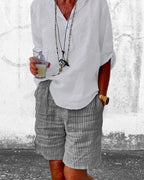 Homme en chemise blanche et short rayé, tenant un verre de boisson. Style décontracté, accessoires tendance, montre dorée. Mode estivale.